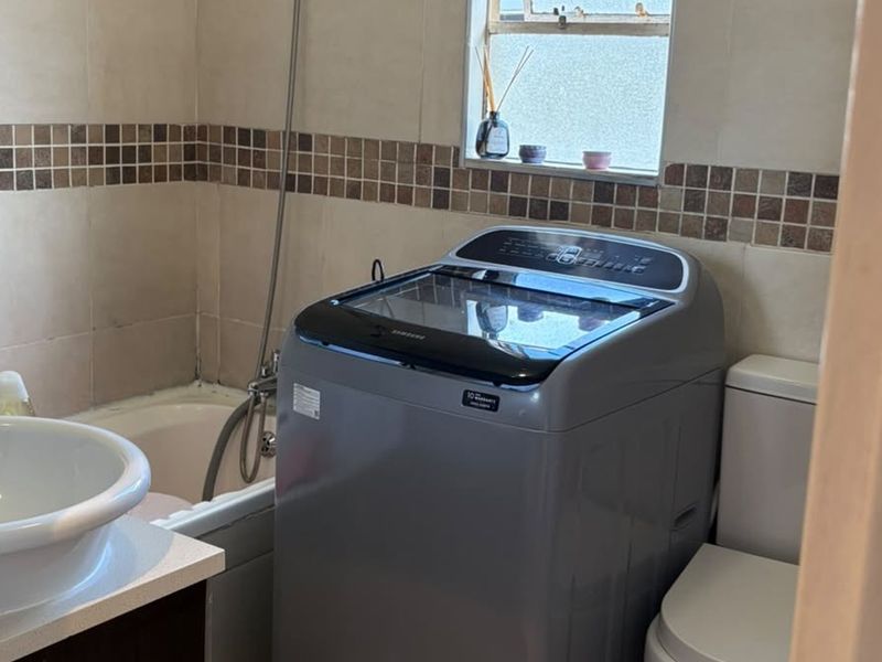 A functional bathroom featuring a bathtub with handheld shower, vanity basin, and toilet, finished with neutral wall tiling and a window for natural light.