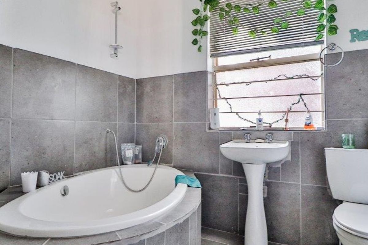 A functional bathroom featuring a built-in bathtub, pedestal sink, and toilet, finished with grey wall tiling and a window with security bars.