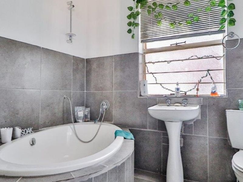 A functional bathroom featuring a built-in bathtub, pedestal sink, and toilet, finished with grey wall tiling and a window with security bars.