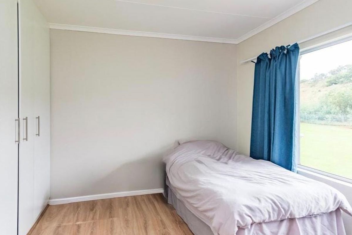 A functional bedroom featuring light-toned laminate flooring, neutral wall finishes, and ample built-in cupboard storage with a large window providing natural light.