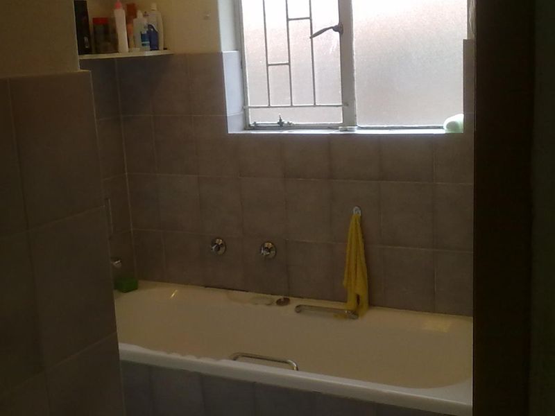 A functional bathroom featuring a standard bathtub with wall-mounted mixer taps and tiled splashbacks, complemented by a window with security bars.