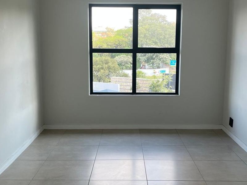 A bright, empty bedroom featuring neutral tiled flooring and a large black aluminium window frame with a view of the surrounding greenery.