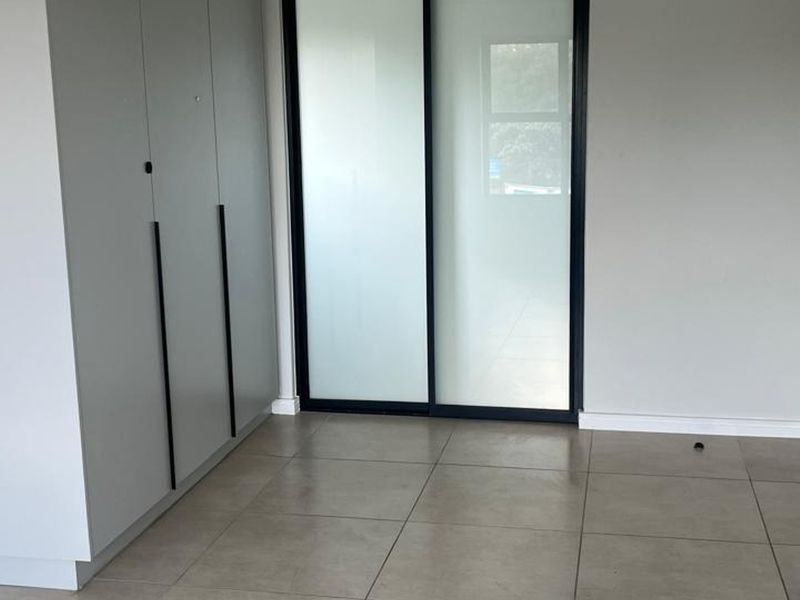 A bright, modern bedroom featuring neutral tiled flooring, built-in cupboards with sleek handles, and a sliding glass door leading to an outdoor area.
