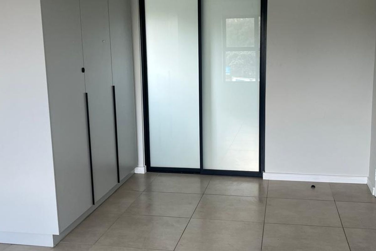 A bright, modern bedroom featuring neutral tiled flooring, built-in cupboards with sleek handles, and a sliding glass door leading to an outdoor area.