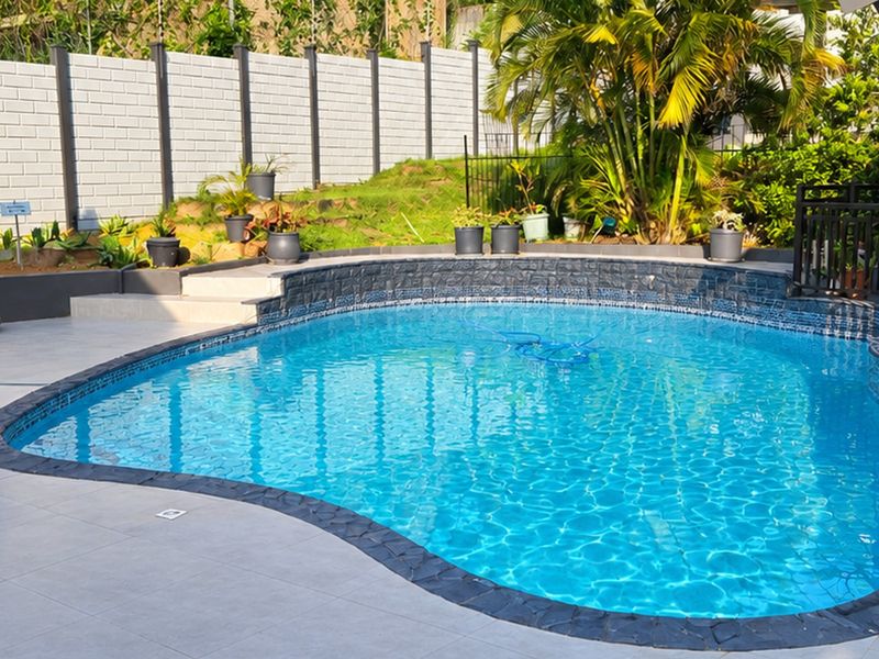 A well-maintained swimming pool area featuring a tiled surround, stone-look pool coping, and a secure boundary wall with landscaped garden backdrop.