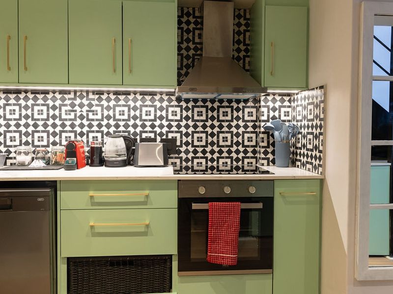 Modern kitchen featuring sage green cabinetry, gold hardware, a striking geometric tile backsplash, stainless steel oven, extractor fan, and integrated dishwasher.