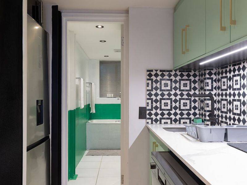 A modern kitchen featuring mint green cabinetry, a striking geometric tile backsplash, and integrated lighting, with a view into the adjacent bathroom.