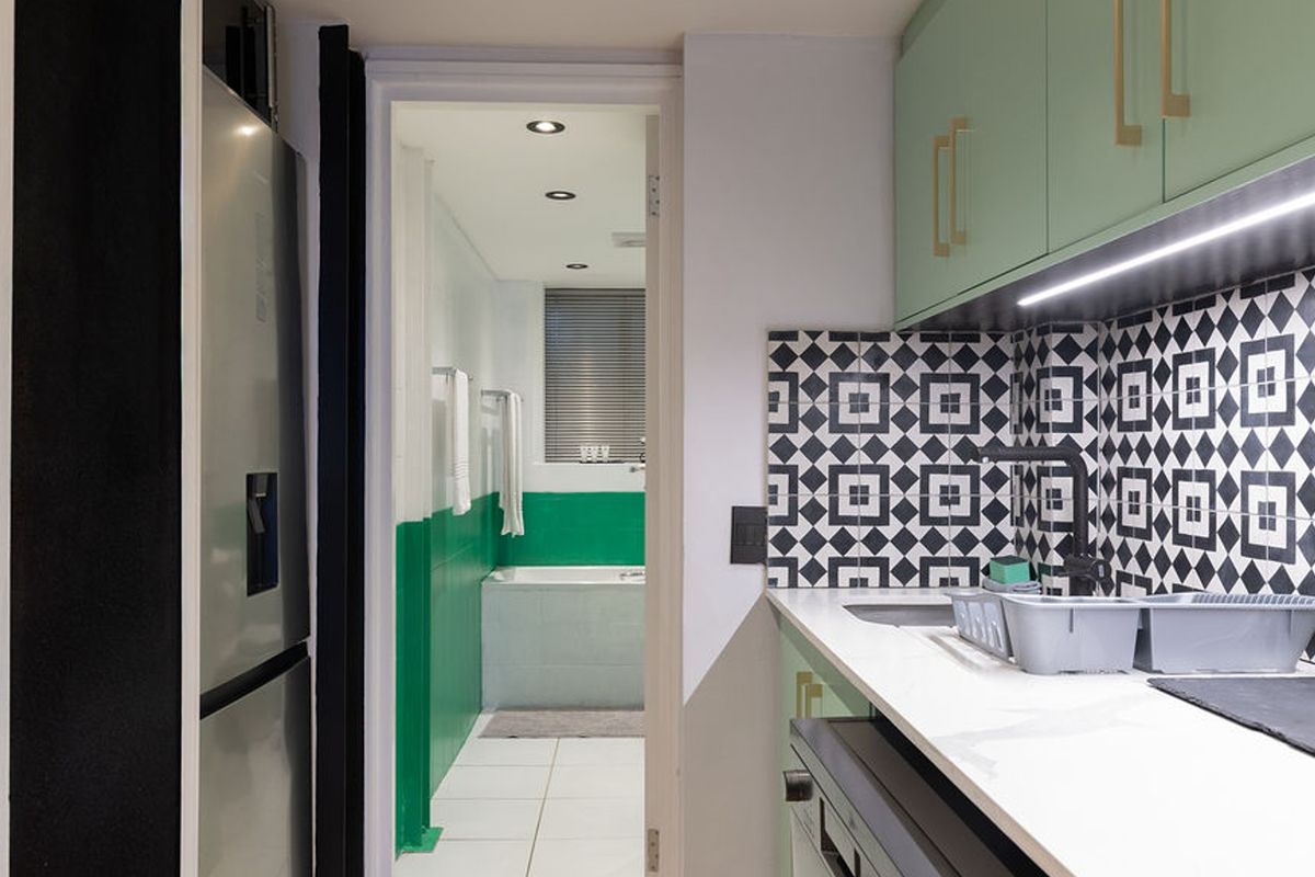A modern kitchen featuring mint green cabinetry, a striking geometric tile backsplash, and integrated lighting, with a view into the adjacent bathroom.