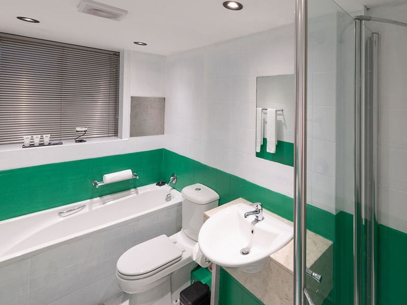 A clean, functional bathroom featuring a bathtub, separate shower enclosure, pedestal basin, and toilet, finished with white and green wall tiling.