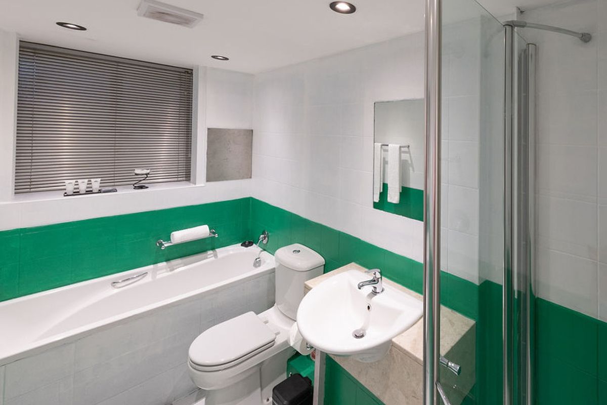 A clean, functional bathroom featuring a bathtub, separate shower enclosure, pedestal basin, and toilet, finished with white and green wall tiling.