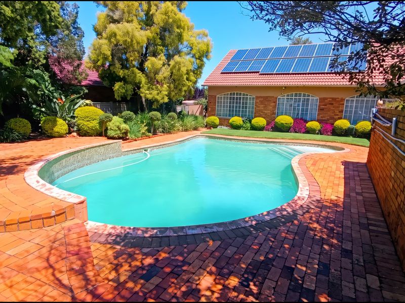 A well-maintained outdoor pool area featuring brick paving, landscaped garden borders, and a house roof equipped with solar panels.