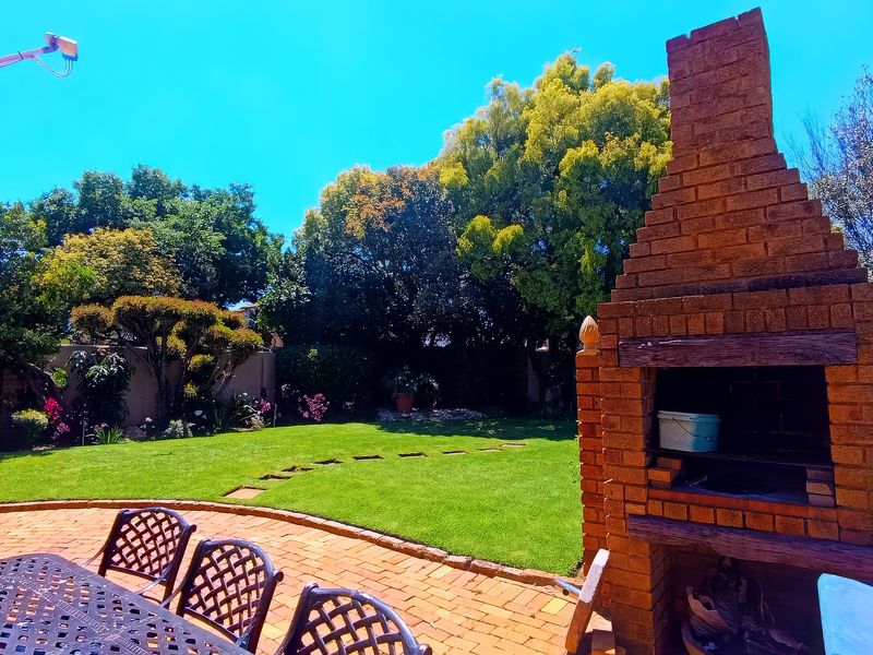 A sunny outdoor patio area featuring a traditional brick built-in braai, brick paving, and a well-maintained garden with mature trees.