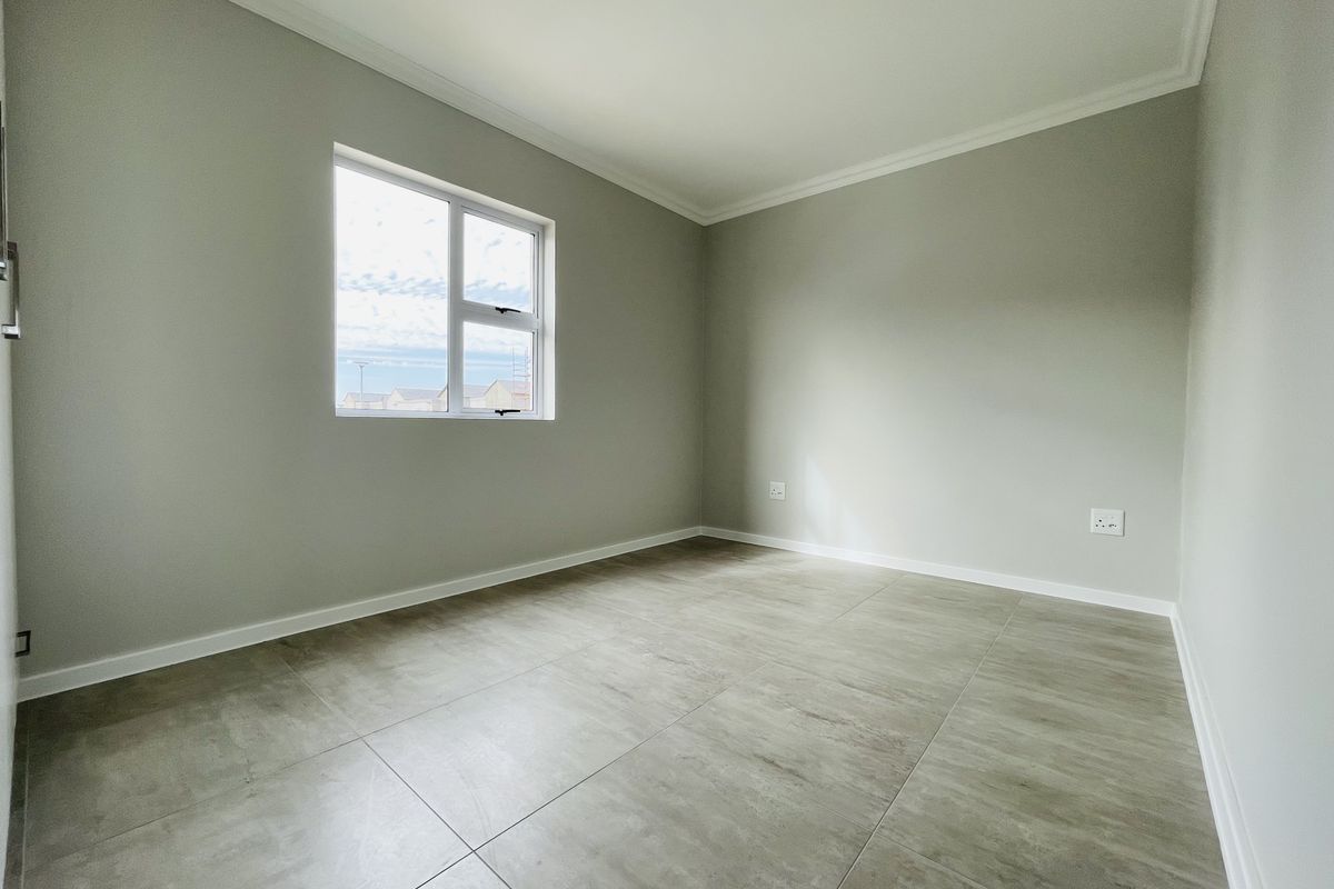 A bright, empty bedroom featuring neutral grey walls, large format floor tiles, and a window providing natural light.