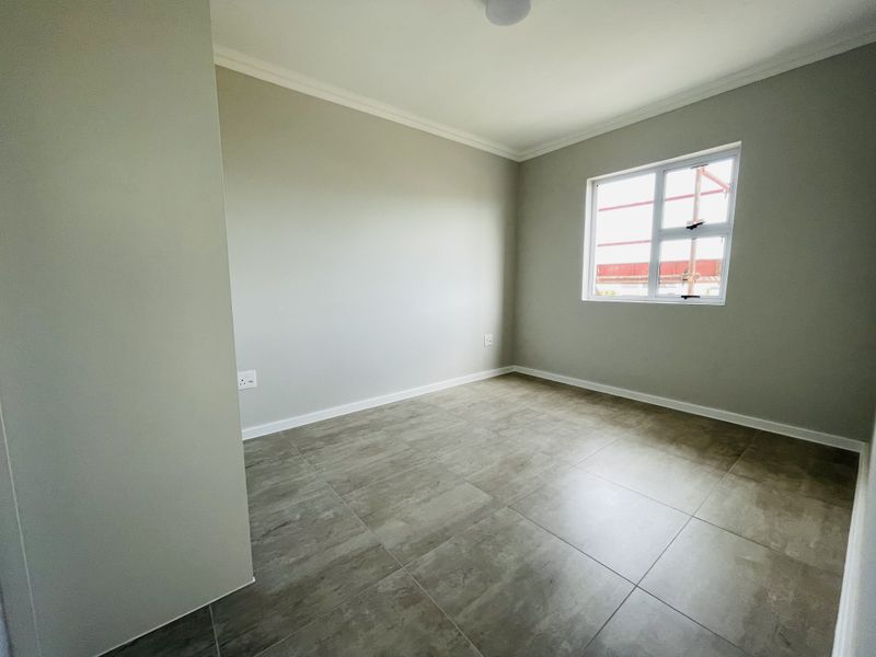 A clean, empty bedroom featuring neutral grey walls, modern large-format floor tiles, and a white-framed window.