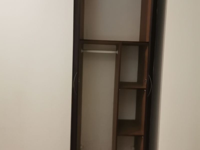 A bedroom featuring a built-in wooden wardrobe with shelving and hanging space, set against plain walls with tiled flooring.