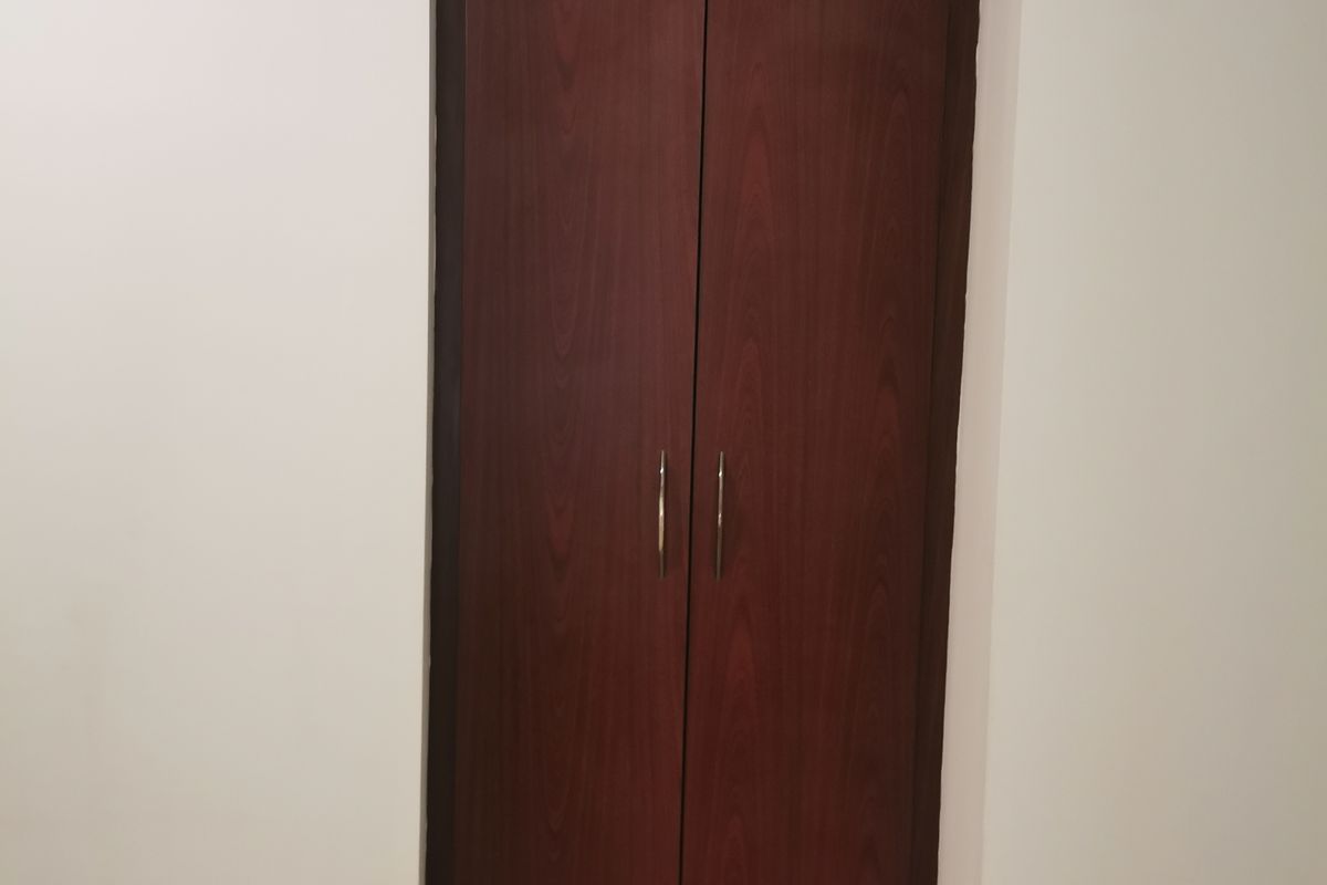 A simple bedroom interior featuring a dark wood built-in cupboard and neutral tiled flooring.