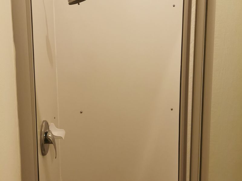 A compact shower stall featuring basic wall panels, a standard shower head, and a mixer tap, showing signs of wear and minor damage.