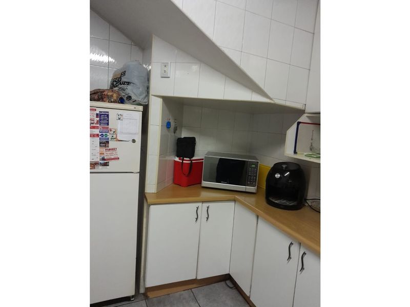 A compact kitchen area featuring white cabinetry, laminate countertops, and tiled splashbacks, situated under a staircase bulkhead.