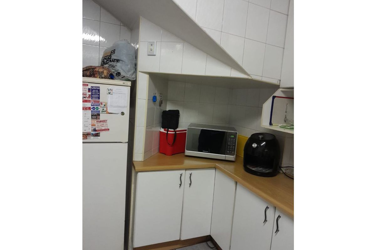 A compact kitchen area featuring white cabinetry, laminate countertops, and tiled splashbacks, situated under a staircase bulkhead.