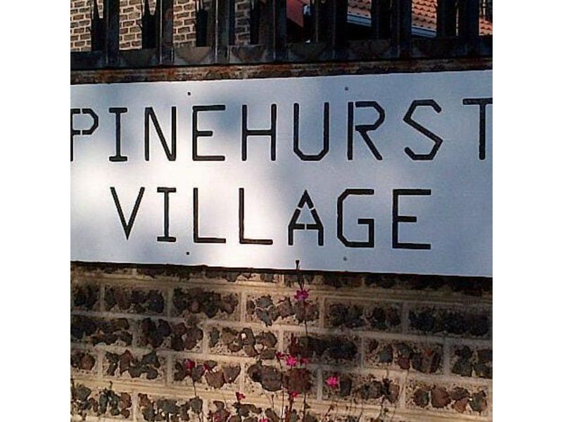A close-up view of the Pinehurst Village signage mounted on a stone masonry wall with metal security fencing above.