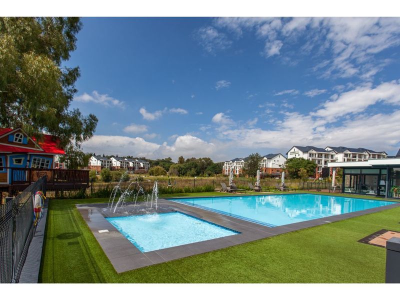 A well-maintained communal swimming pool area featuring a wading pool with water fountains, surrounded by artificial turf and modern apartment complex buildings.