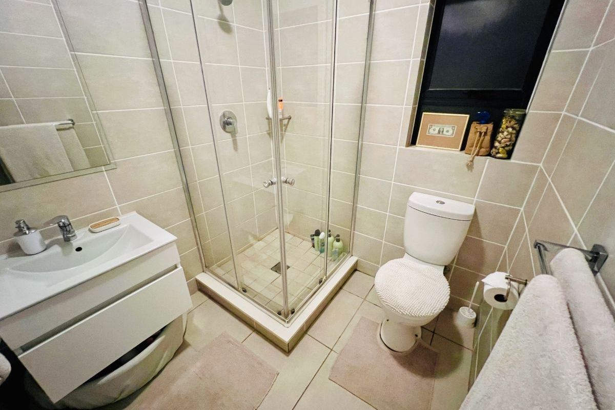 A compact, modern bathroom featuring a glass shower enclosure, wall-mounted vanity with basin, and a standard toilet with neutral wall and floor tiling.