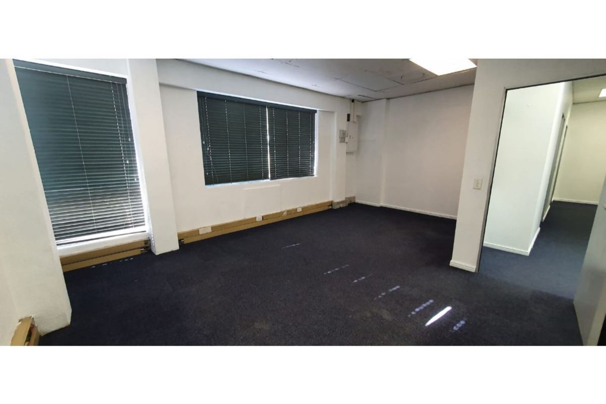 An empty, open-plan office space featuring dark carpeting, large windows with venetian blinds, and a suspended ceiling with recessed lighting.