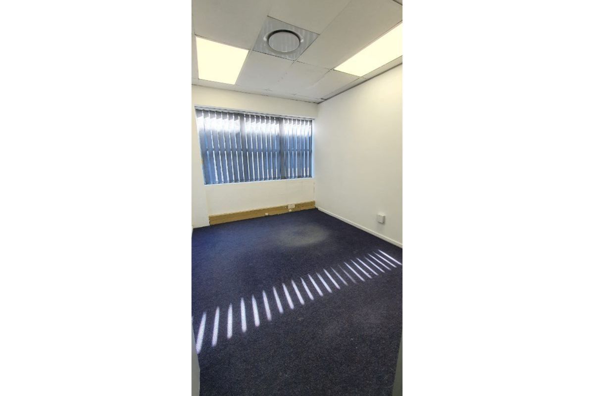 A vacant office space featuring blue carpeting, a suspended ceiling with recessed lighting, and vertical window blinds.