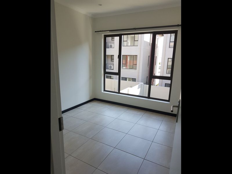 A bright, empty bedroom featuring neutral tiled flooring and a large window providing natural light and views of the surrounding apartment complex.