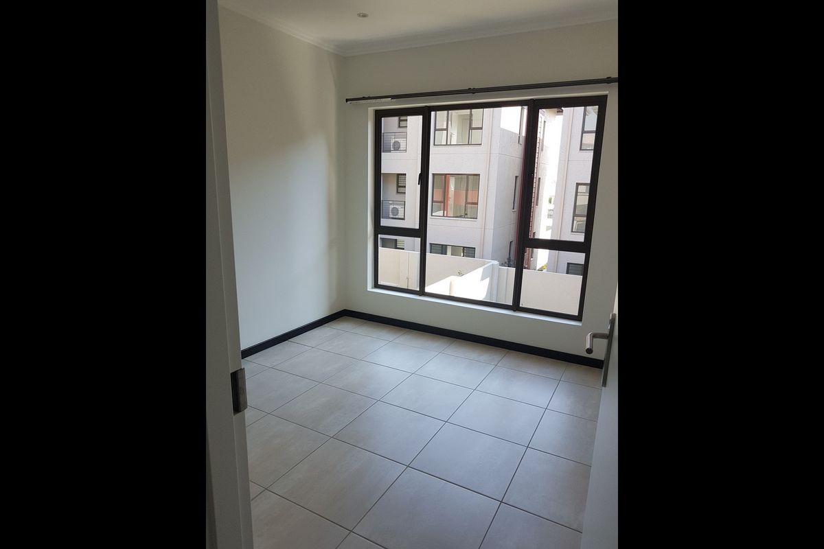 A bright, empty bedroom featuring neutral tiled flooring and a large window providing natural light and views of the surrounding apartment complex.