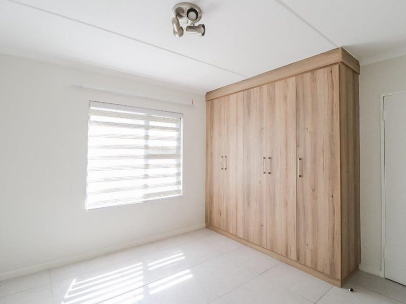 A bright, empty bedroom featuring modern light wood-finish built-in cupboards, tiled flooring, and fitted window blinds.
