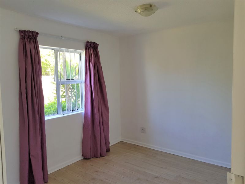 A bright, empty bedroom featuring light-toned laminate flooring, a window with security bars, and neutral wall finishes.