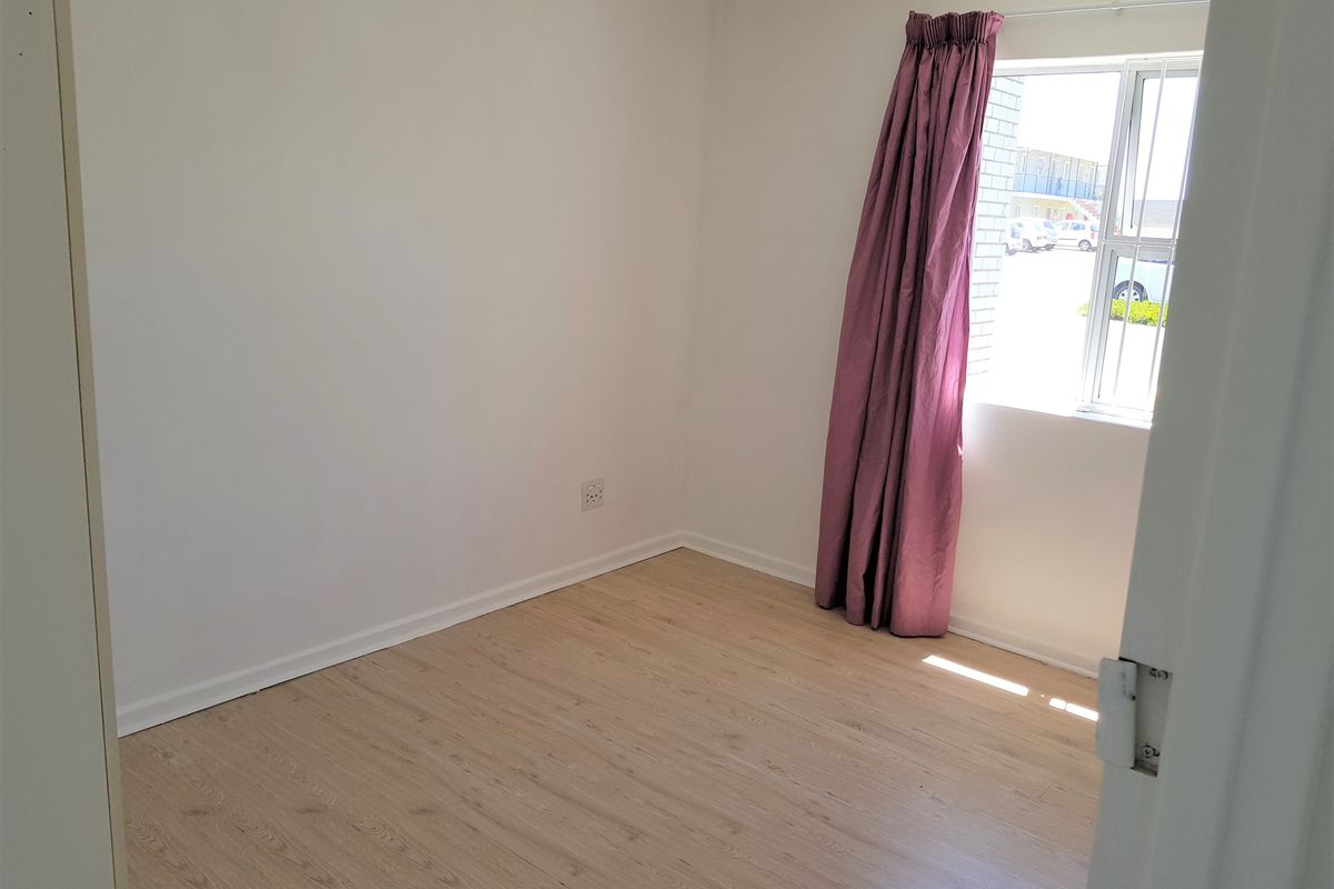 A bright, empty bedroom featuring light-toned laminate flooring, white walls, and a window fitted with security bars.