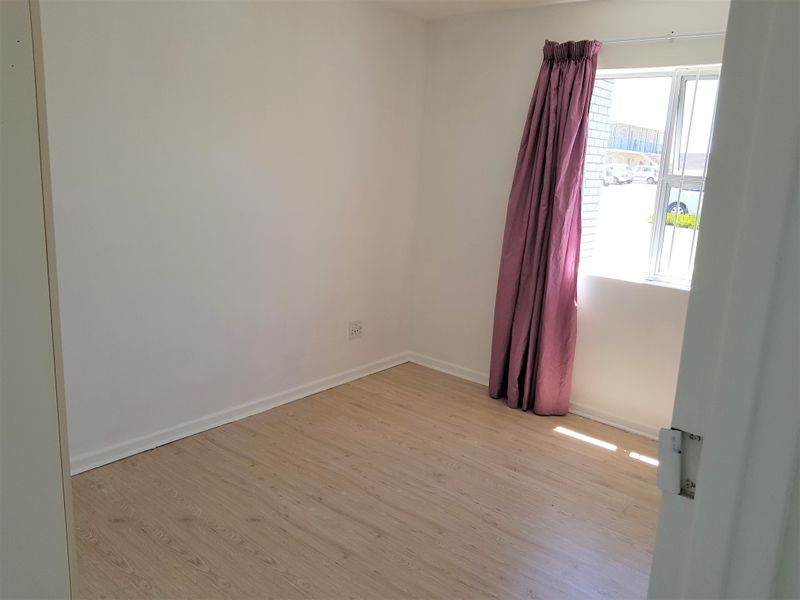 A bright, empty bedroom featuring light-toned laminate flooring, white walls, and a window fitted with security bars.