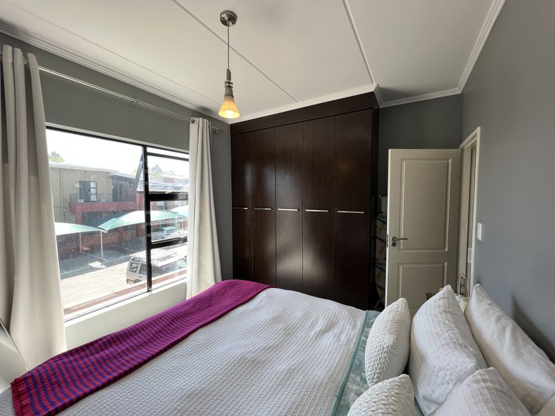 A well-maintained bedroom featuring dark wood built-in cupboards, a modern pendant light, and large windows providing natural light and views of the complex.