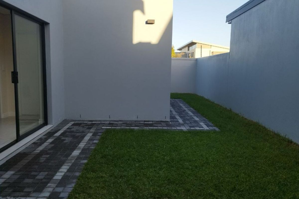A private rear garden area featuring a paved walkway, manicured lawn, and a sliding glass door providing access to the interior.