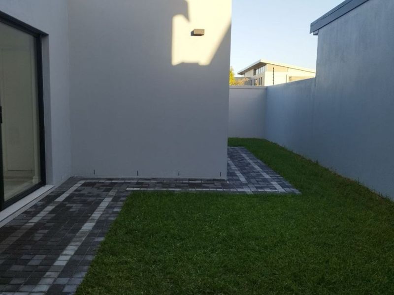 A private rear garden area featuring a paved walkway, manicured lawn, and a sliding glass door providing access to the interior.
