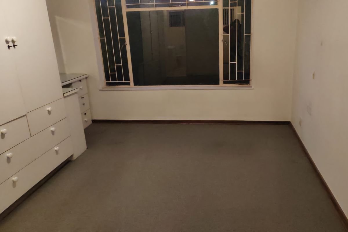 A vacant bedroom featuring built-in cupboards and a window with security bars, requiring cosmetic updates to the flooring and wall finishes.