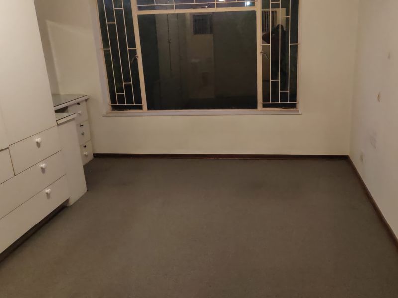 A vacant bedroom featuring built-in cupboards and a window with security bars, requiring cosmetic updates to the flooring and wall finishes.