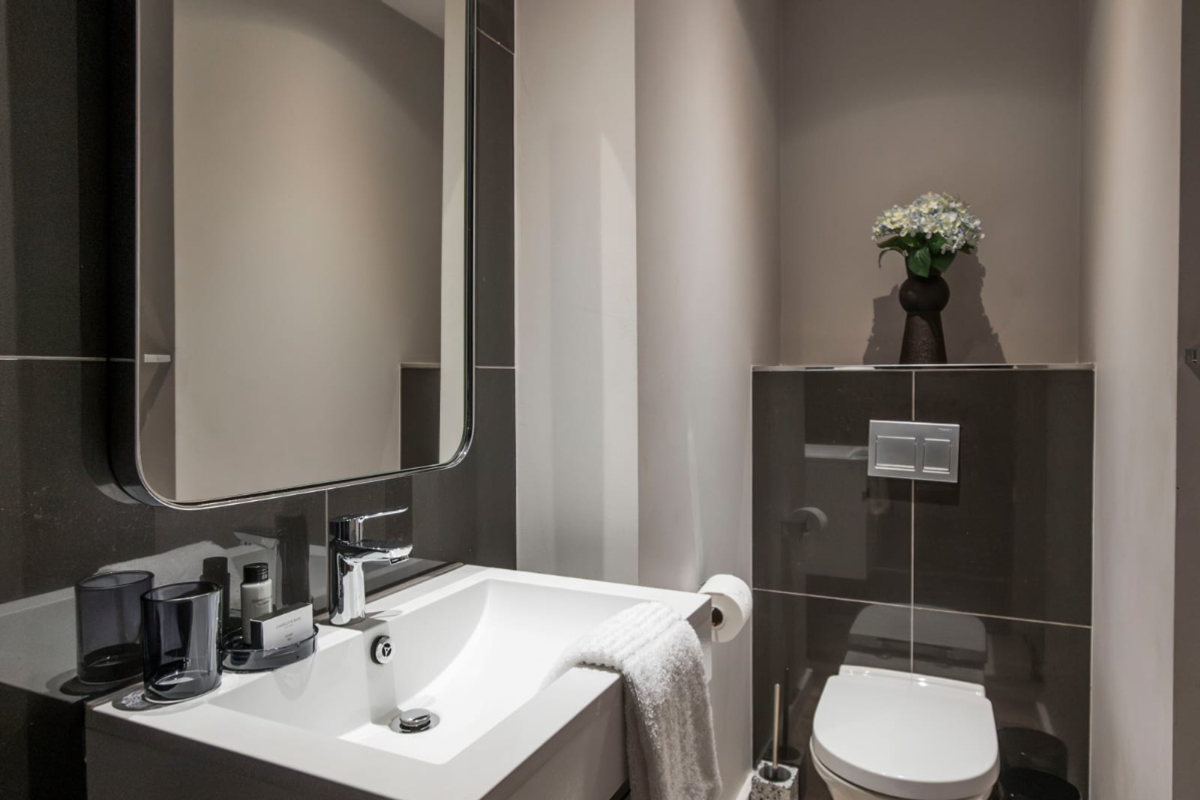 A modern, well-appointed bathroom featuring a wall-mounted toilet, sleek integrated vanity basin with chrome fixtures, and large wall mirror with dark tiled accents.