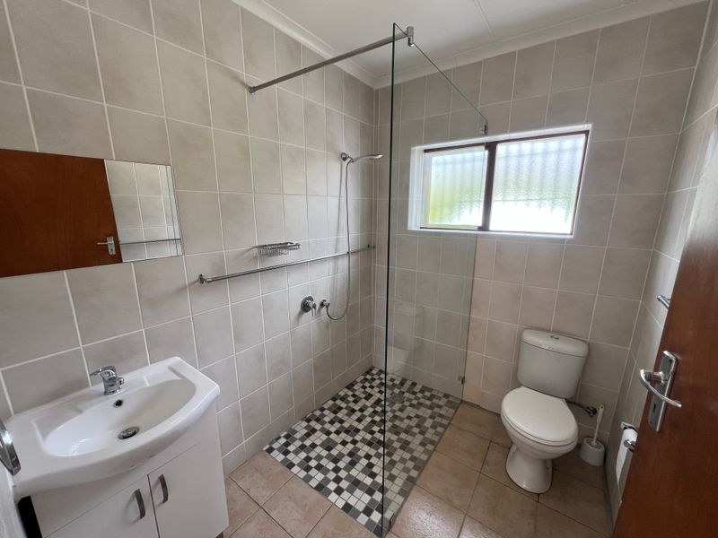 A clean, functional bathroom featuring a walk-in shower with a glass screen, vanity basin, and toilet, finished with neutral wall and floor tiling.