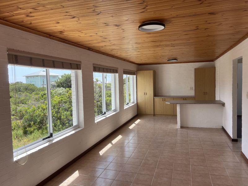 Bright open-plan living area featuring a timber ceiling, tiled floors, and built-in cabinetry with a breakfast bar, offering pleasant views through large windows.