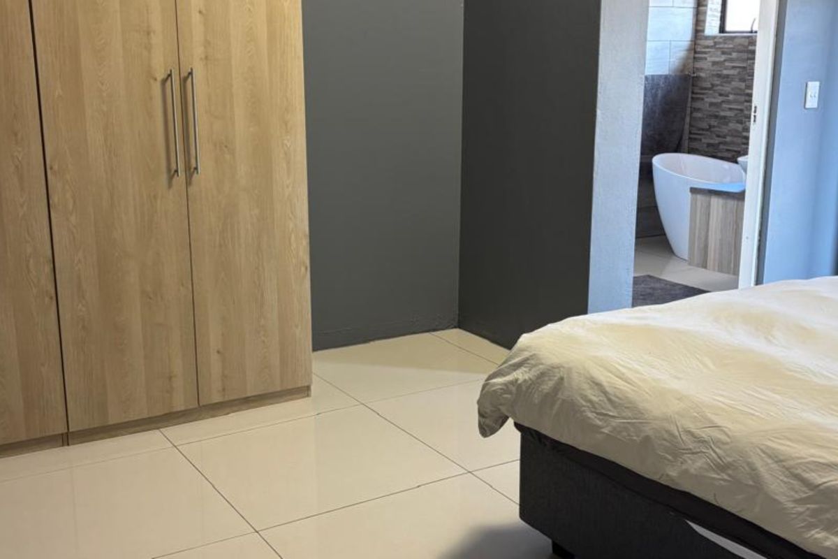 A modern bedroom featuring light wood built-in cupboards, neutral tiled flooring, and direct access to an en-suite bathroom.