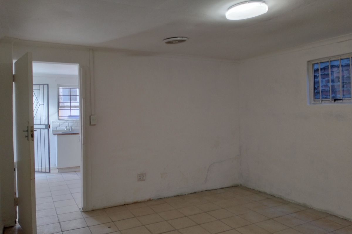 An empty, neutrally painted living area with tiled flooring and a view into an adjacent kitchen space.