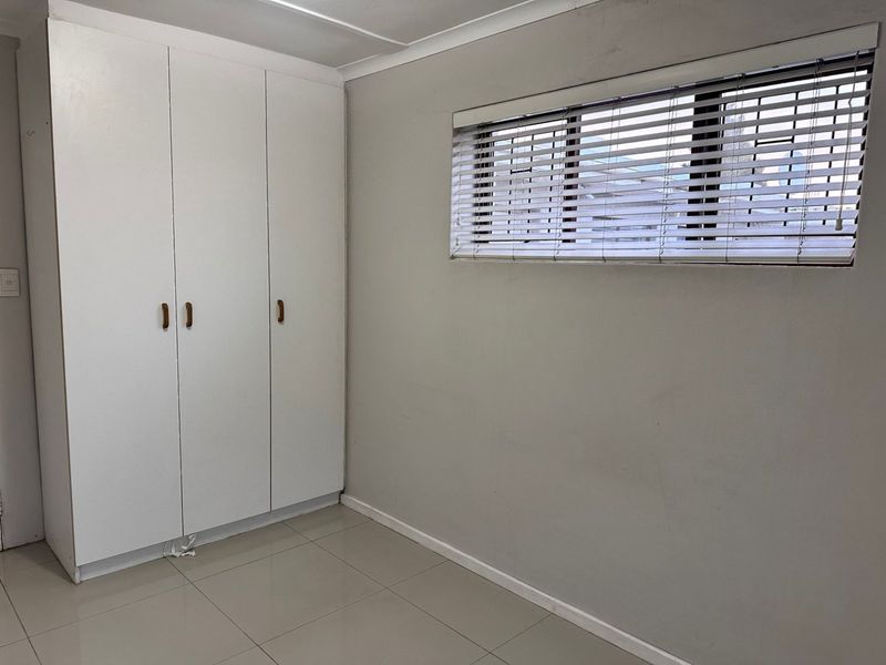A clean, empty bedroom featuring neutral tiled flooring, built-in white cupboards, and a window fitted with venetian blinds.