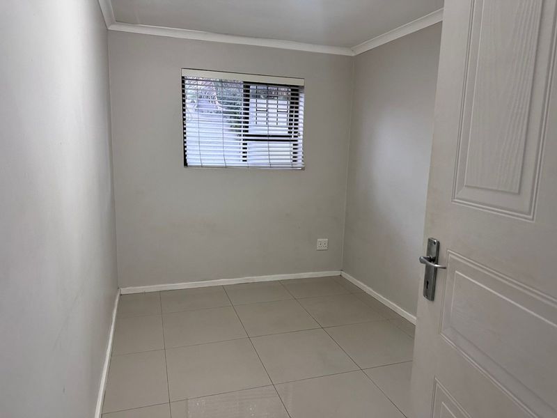 A clean, empty bedroom featuring neutral tiled flooring, light grey walls, and a window fitted with venetian blinds.