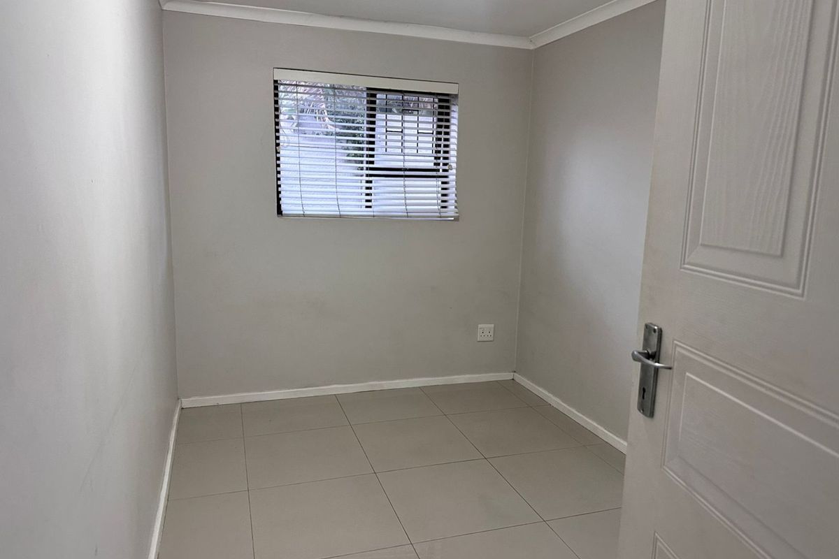 A clean, empty bedroom featuring neutral tiled flooring, light grey walls, and a window fitted with venetian blinds.