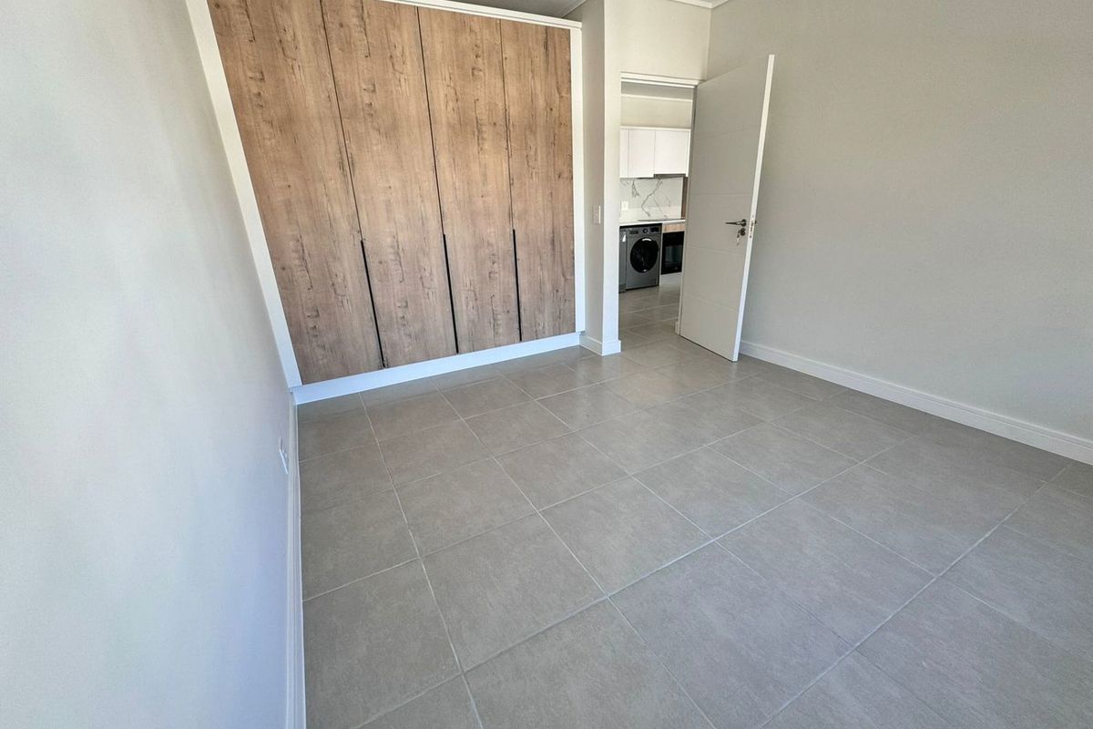A bright, modern bedroom featuring neutral floor tiles and built-in wooden cabinetry, with a view into the adjacent kitchen area.