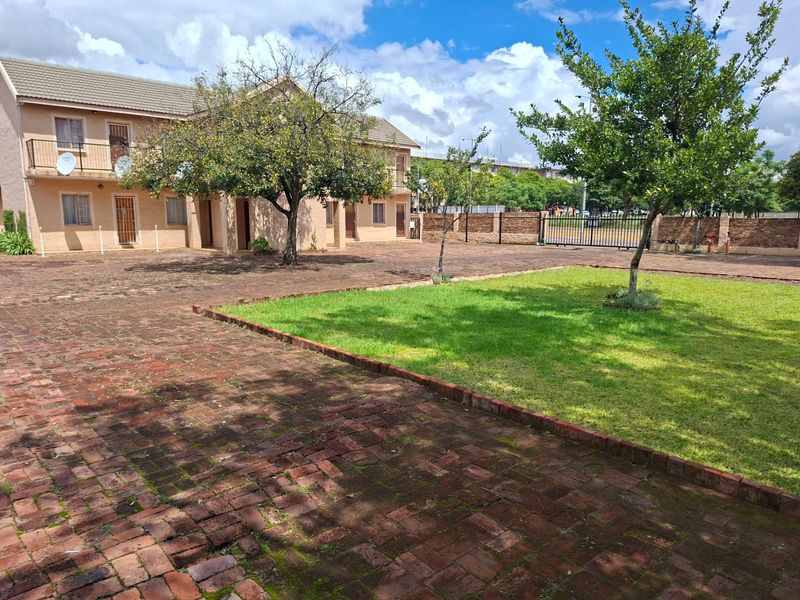 A view of the rear exterior area featuring a brick-paved courtyard, a well-maintained lawn, and a secure perimeter wall with a metal gate.
