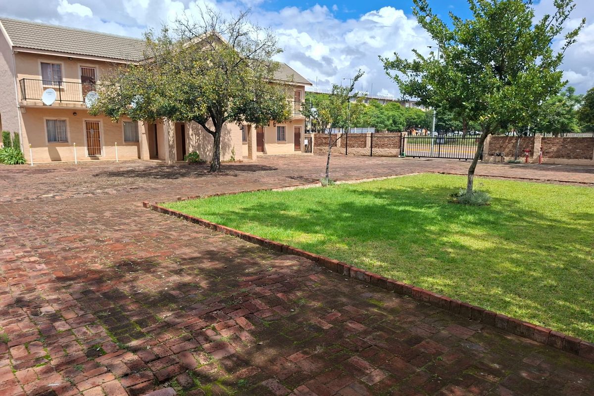 A view of the rear exterior area featuring a brick-paved courtyard, a well-maintained lawn, and a secure perimeter wall with a metal gate.